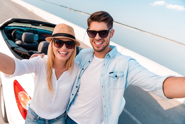 Capturing the bright moments. Top view of cheerful young couple making selfie while standing near their white convertible