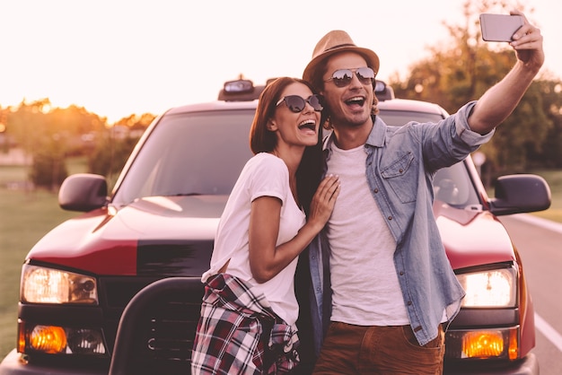 Capturing fun. Beautiful young couple bonding to each other and leaning at their pick-up truck while making selfie