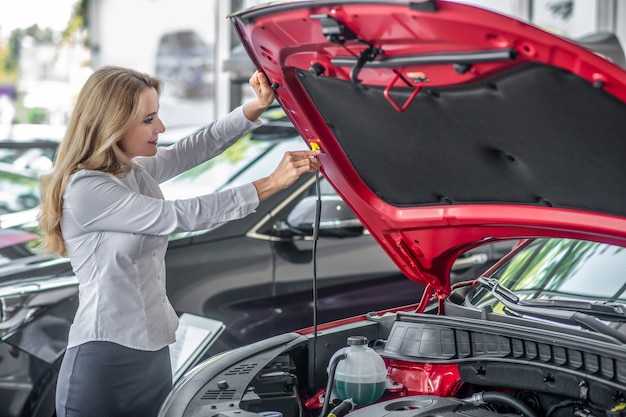 Car bonnet. Smiling confident blonde woman in white blouse opening hood of new red car in car dealership in afternoon