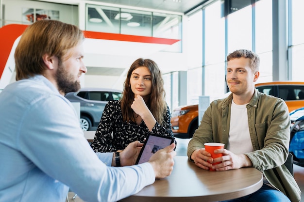 A car dealership manager advises a young couple before buying a new family car at a dealership A man and a young woman drink coffee and listen to the manager