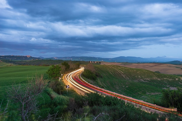 Car light trail on winding road in the unique hill landscape of Tuscany Italy Dramatic sunset sky at twilight illuminated villages on hilltop