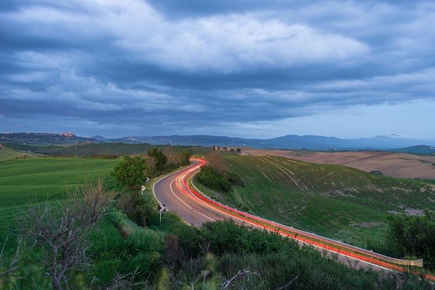 Car light trail on winding road in the unique hill landscape of Tuscany Italy Dramatic sunset sky at twilight illuminated villages on hilltop