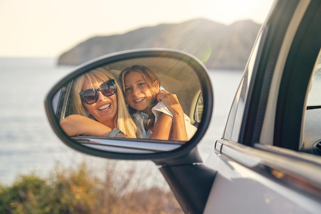 Car mirror with the image of a blond mother and daughter hugging and looking at the mirror in front of the sea