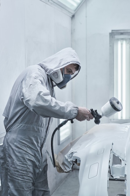 Car painting and automobile repair service. Auto mechanic in white overalls paints car with airbrush pulverizer in paint chamber