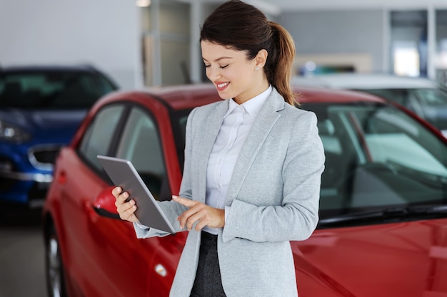 Car seller in suit standing in car salon and using tablet for choosing right car.