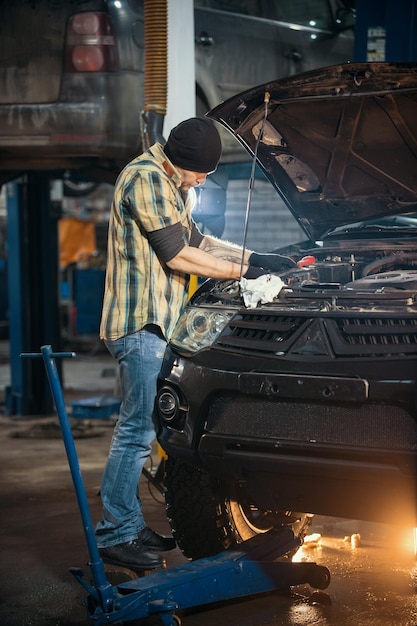 Car service brutal mechanic man standing by the car with open hood and inspects the engine