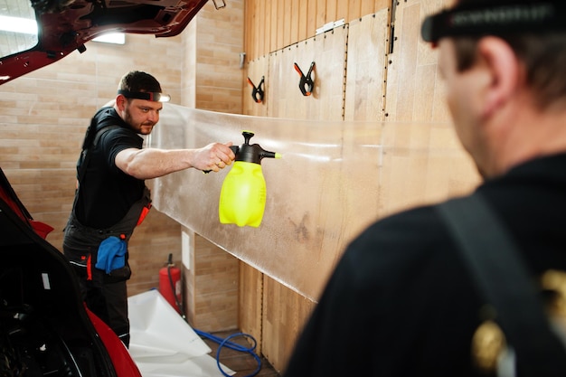 Car service worker put anti gravel film on a red car body at the detailing vehicle workshop Car protection with special films