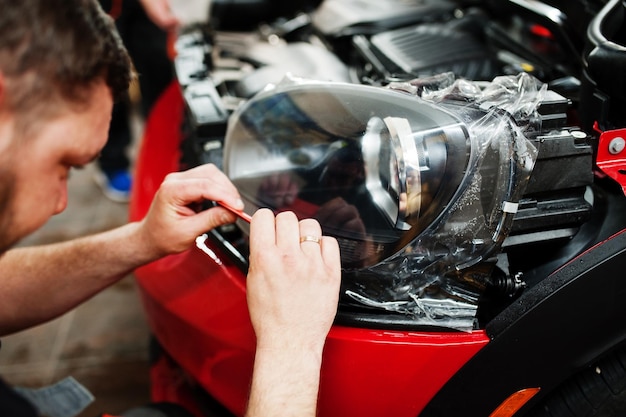 Car service worker put anti gravel film on a red car body at the detailing vehicle workshop Car protection with special films