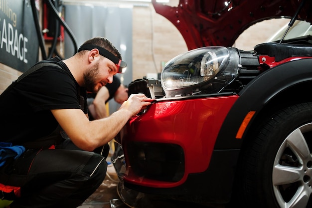 Car service worker put anti gravel film on a red car body at the detailing vehicle workshop Car protection with special films