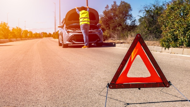 Car with problems and a red triangle to warn other road users Male driver standing near a broken car with open up hood