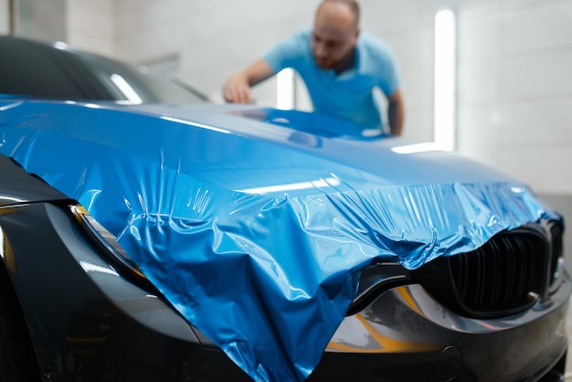 Car wrapping, man installs protective vinyl foil or film on hood. Worker makes auto detailing