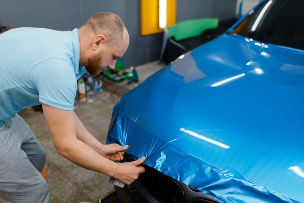 Car wrapping, man with squeegee installs protective vinyl foil or film on hood. Worker makes auto detailing