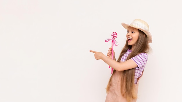 A caramel lollipop in the hands of a beautiful little girl A baby in a summer sundress and a summer hat points to a white isolated background with her index finger