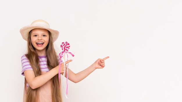 A caramel lollipop in the hands of a beautiful little girl A baby in a summer sundress and a summer hat points to a white isolated background with her index finger