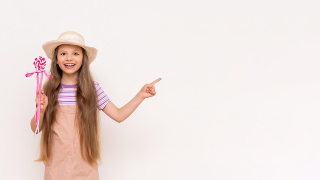 A caramel lollipop in the hands of a beautiful little girl A baby in a summer sundress and a summer hat points to a white isolated background with her index finger