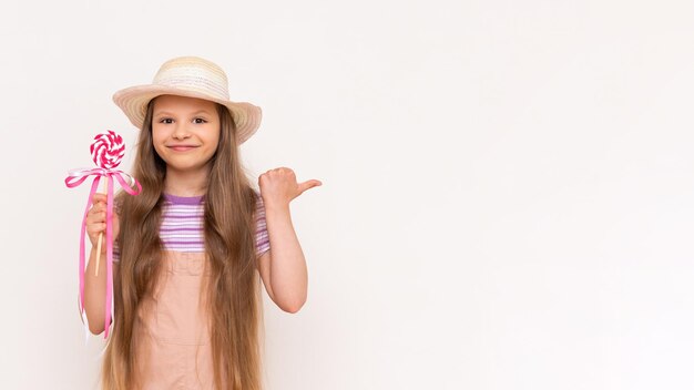 A caramel lollipop in the hands of a beautiful little girl A little girl in a summer sundress and a summer hat points with her thumb at a white isolated background