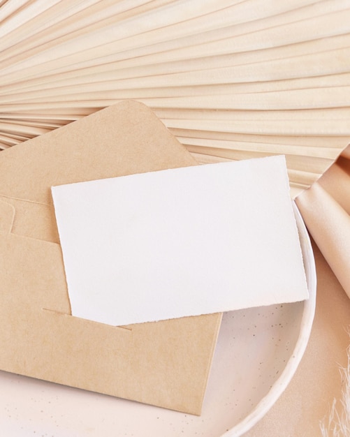 Card and envelope on plate on dried palm leaf and beige silky fabric close up greeting or wedding mockup