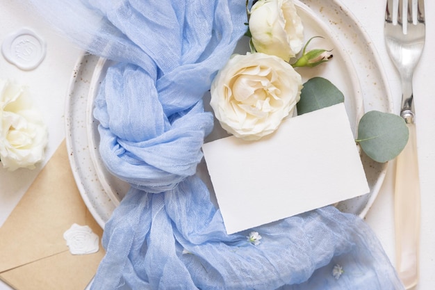 Card near light blue tulle fabric knot and cream roses on plates top view copy space wedding mockup