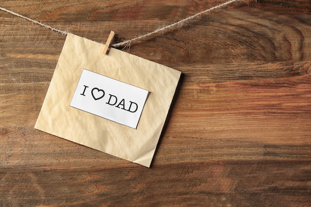 Card with text I LOVE DAD hanging on rope against wooden background