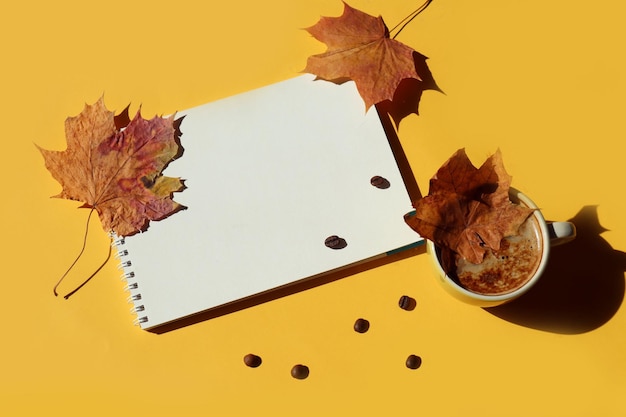 A card with the wishes of a cozy autumn a notebook with maple leaves on it a cup with a maple leaf next to it scattered coffee beans a yellow background a place for text