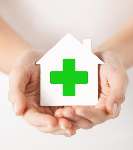 care, help, charity and people concept - close up of hands holding white paper house with green cross sign