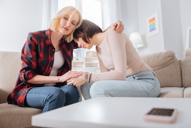 Care and support. Nice pleasant attractive woman sitting on the sofa and hugging her girlfriend while expressing her support