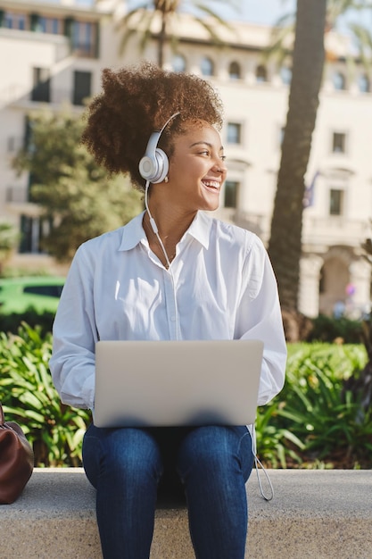 Carefree African American female freelancer in casual clothes sitting on stone bench in city park and working remotely on laptop while listening to music in headphones