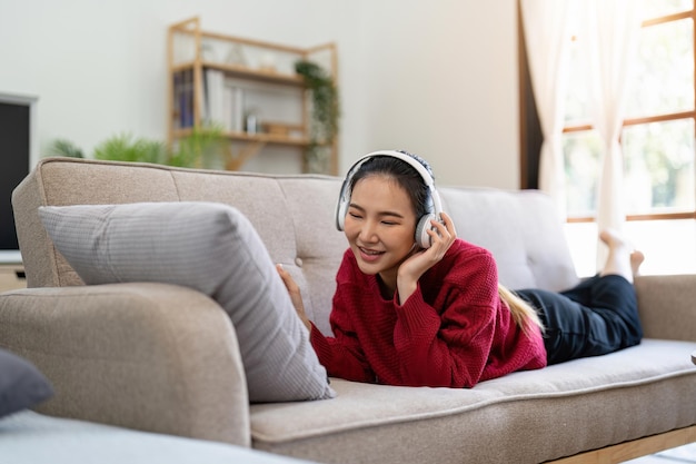 Carefree asian woman listening to music using headphones laying on couch in living room copy space Happy young lady with closed eyes enjoying music home interior