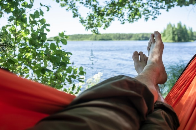 Carefree barefoot man relaxes lying in hammock in shade of trees with a magnificent view of the lake