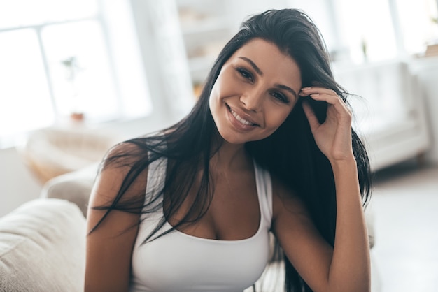 Carefree beauty. Portrait of attractive young woman adjusting her hair and smiling while sitting on the couch at home