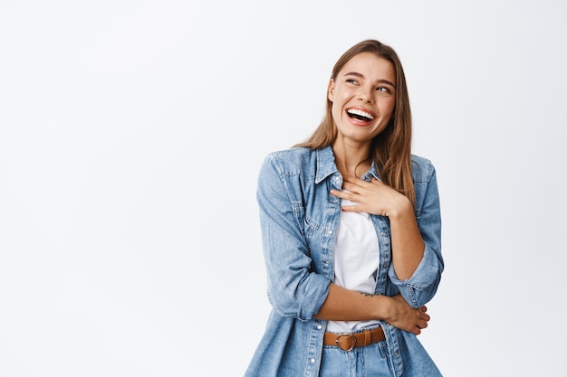 Carefree blond girl laughing and looking canid, holding hand on chest and chuckling about something funny, staring left at copy space banner, white