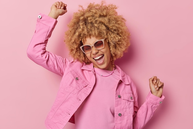 Carefree cheerful European woman with curly blonde hair has fun dances and moves with rhythm of music laughs happily wears fashionable jacket trendy sunglasses isolated over pink background