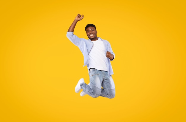 Carefree excited black man in casual outfit jumping in the air and smiling over yellow studio background copy space