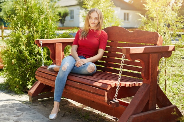 Carefree female with straight hair wearing red sweater and trendy jeans sitting at comfortable wooden bench in park enjoying sunshine