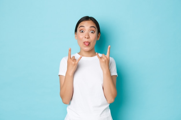 Carefree funny and upbeat pretty asian girl in white t-shirt, showing tongue and rock-n-roll gesture, enjoying awesome festival, looking pleased and amazed, partying over blue background