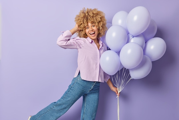 Carefree glad curly haired woman dances with bunch of inflated balloons wears shirt and jeans enjoys party time isolated over purple background People celebration and special occasion concept