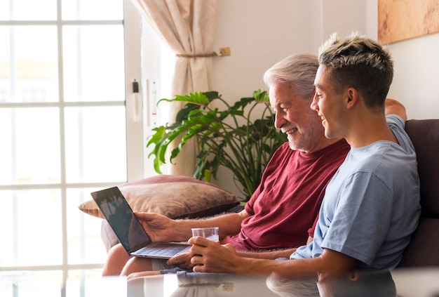 Carefree grandfather and grandson sitting together on the sofa at home looking at the same laptop smiling - sharing the same interest or passion. Concept of family united and friendship