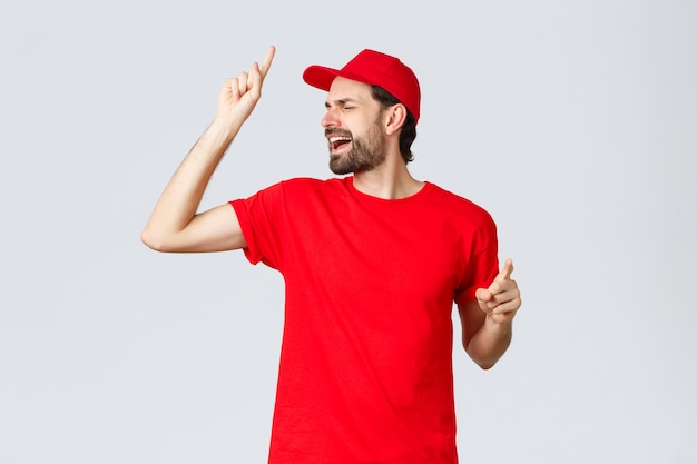 Carefree handsome bearded guy delivery employee in red tshirt and cap singing with closed eyes raisi...