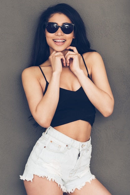 Carefree style. Beautiful young woman in sunglasses looking at camera and smiling while standing against grey background