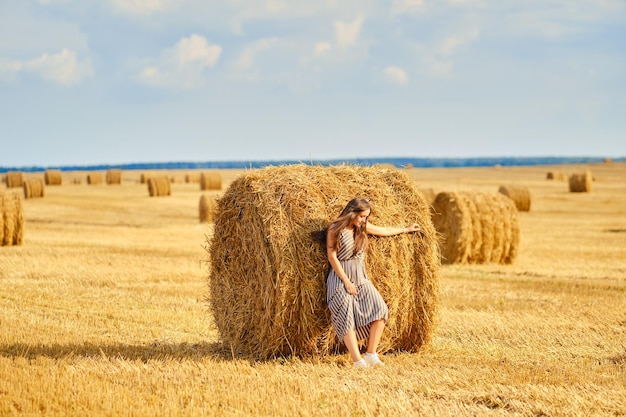 Carefree woman leaning to huge rick of straw on the field in sunny day