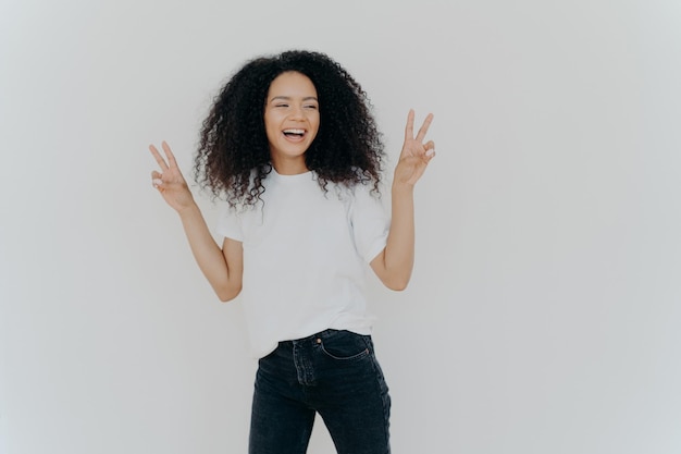 Carefree woman with Afro hairstyle raises hands up shows victory gesture or peace sign looks gladfully aside dressed in casual wear smiles relaxed sends hello isolated over white background