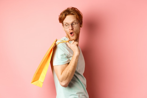 Carefree young man with red hair and glasses, walking with shopping bag, looking behind his shoulder with amazed expression, standing over pink background.