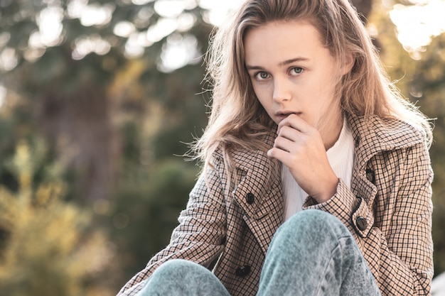 Carefree young woman in trendy vintage pants and coat sitting in park. Cute blonde girl in good mood posing in autumn day, enjoying good weather.