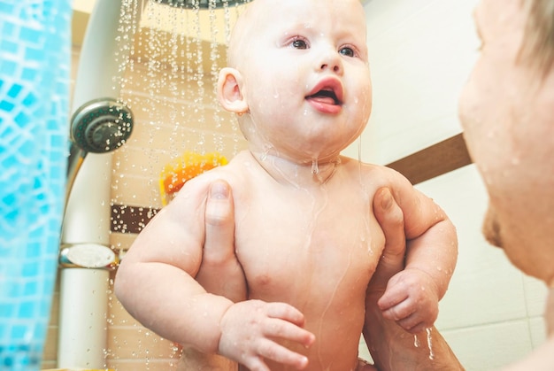 Careful father holds little baby under running shower water in light bathroom at home closeup