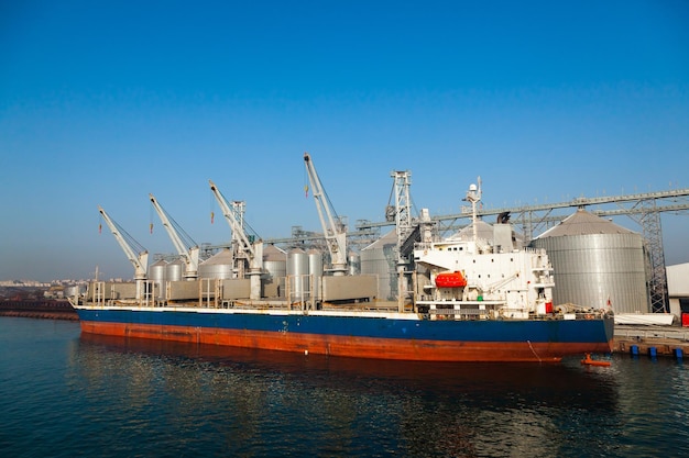 Cargo transport ship moored at grain storage terminal with storage tanks loading lines at seaport