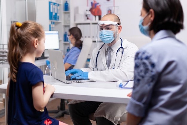 Caring doctor discussing symptoms with mother in medical clinic. Specialist in pediatry medicine with protection mask and visor against covid19 talking about diagnostic and healtcare treatment