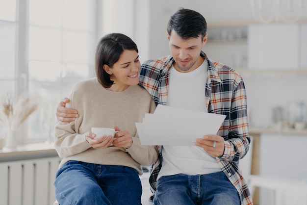 Caring man embraces his wife, look through documents with happy expressions, study payment bills, drinks coffee, dressed in casual wear, pose in spacious room with blurred background. Family and work