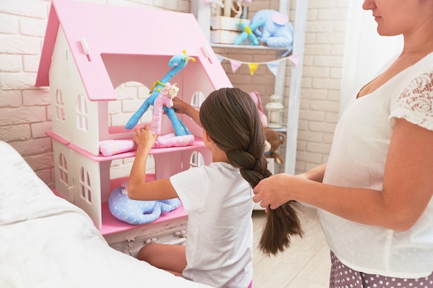 Caring mother braids her daughter's hair when the girl is playing with toys