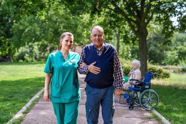 Caring nurse supports senior man walking outdoors with elder in wheelchair in background