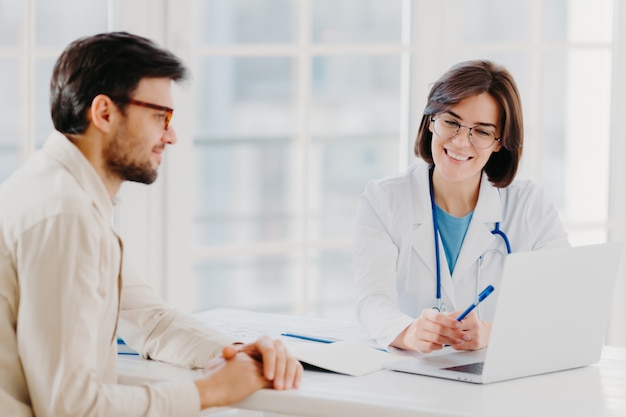 Caring woman doctor gives prescriptions to patient, sit together at table and look at laptop computer